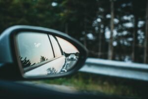 A car is reflected in a side mirror on a scenic forest road.
